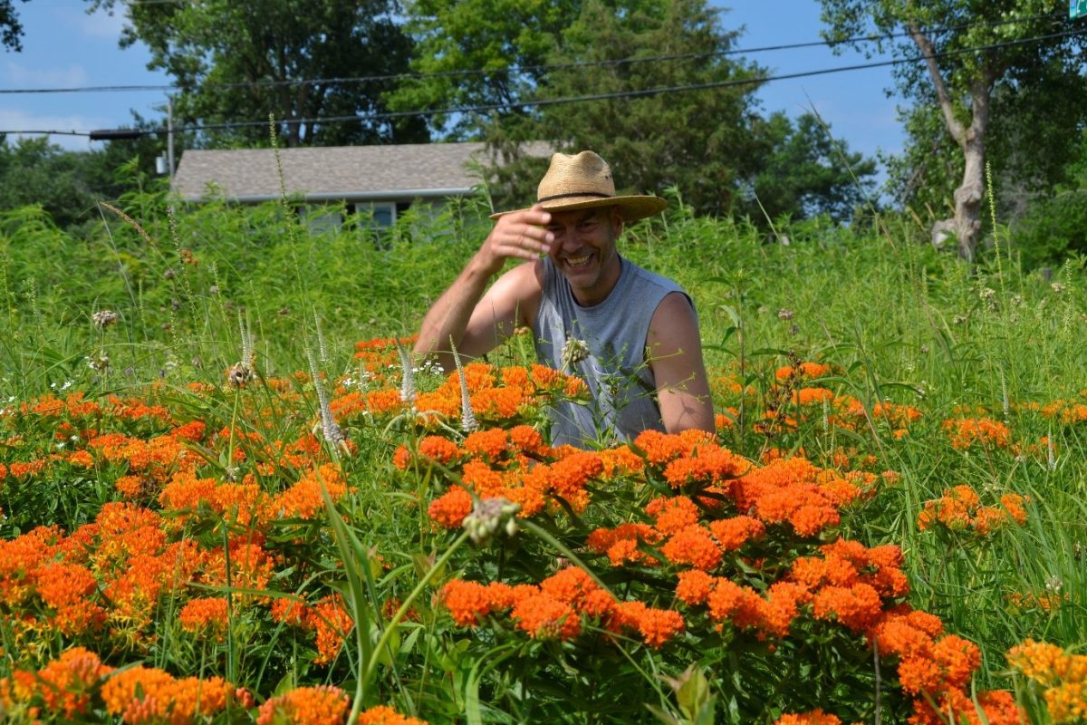 A man in a straw hat sits behind some tall orange flowers looking happy in a field outdoors