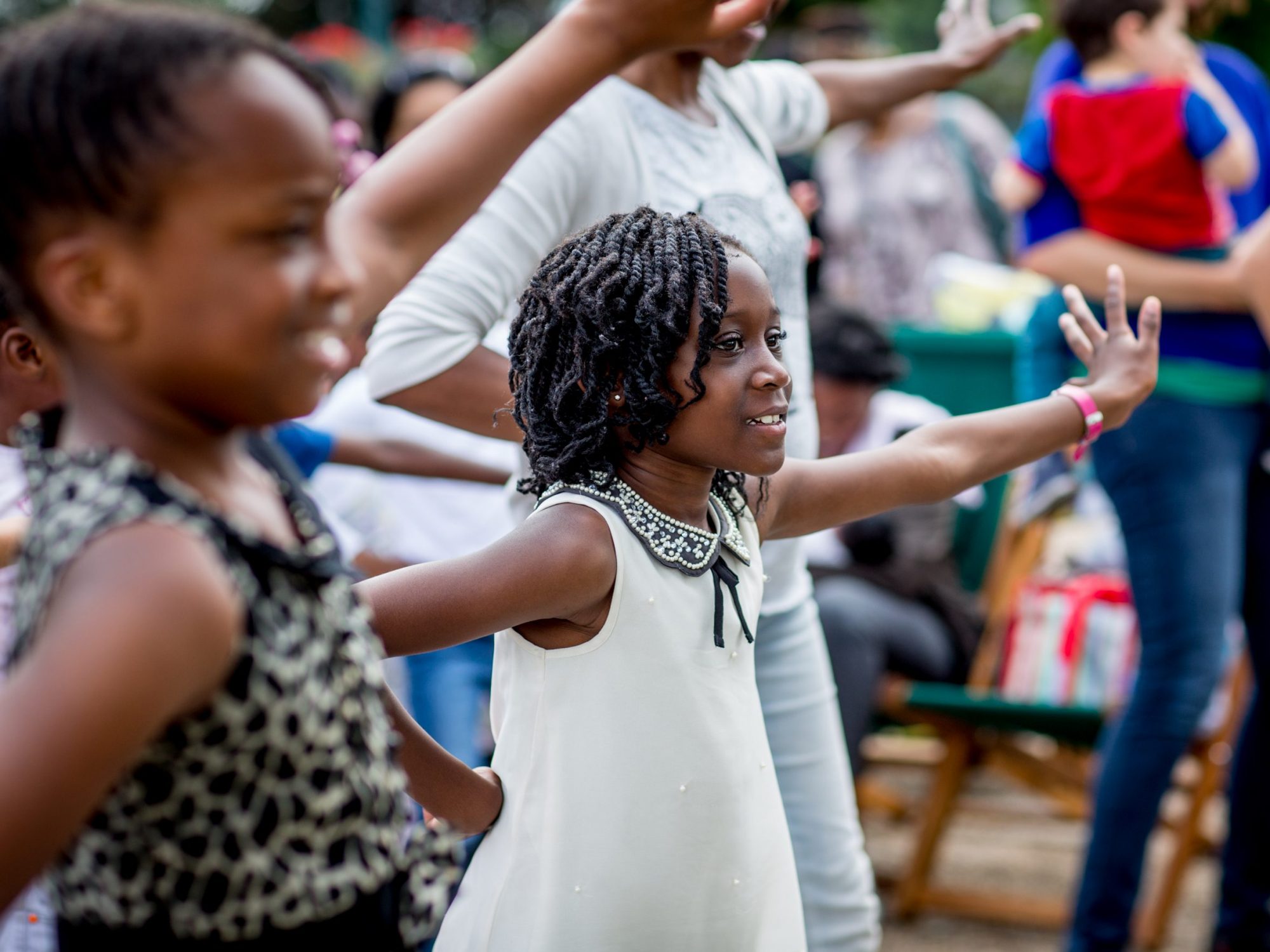 Two girls in close up, in the middle of doing a dance pose, in among other adults and children dancing - they are smiling and their arms are in the air