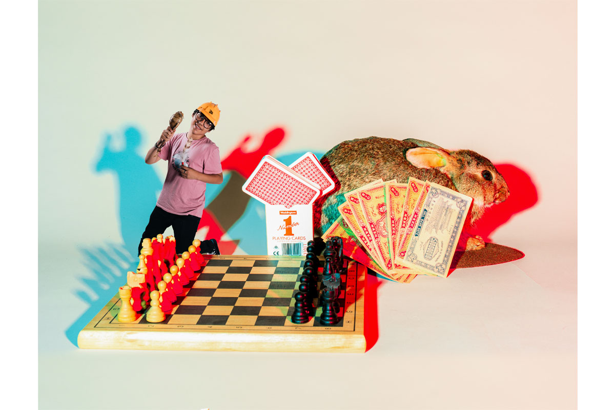 Photo of a young man edited to look small next to a chess board, a deck of cards and a taxidermy rabbit