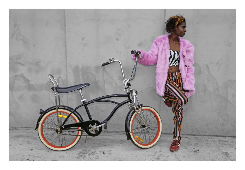 Girl wearing a pink coat holds onto a bike and leans against a wall. She and the bike are in colour and the rest of the image is in black and white.