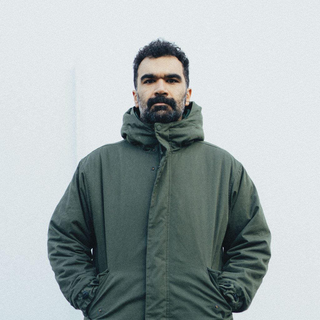 Pouya Eshaei, a man with a beard and moustache, stands in front of a white wall wearing a parka